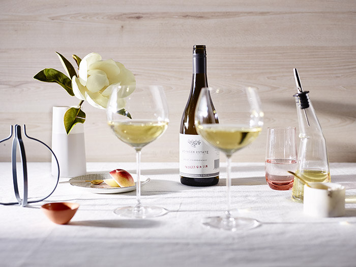 Still life arrangement featuring a bottle of white wine, two glasses filled with white wine, and various decorative objects on a white tablecloth.
