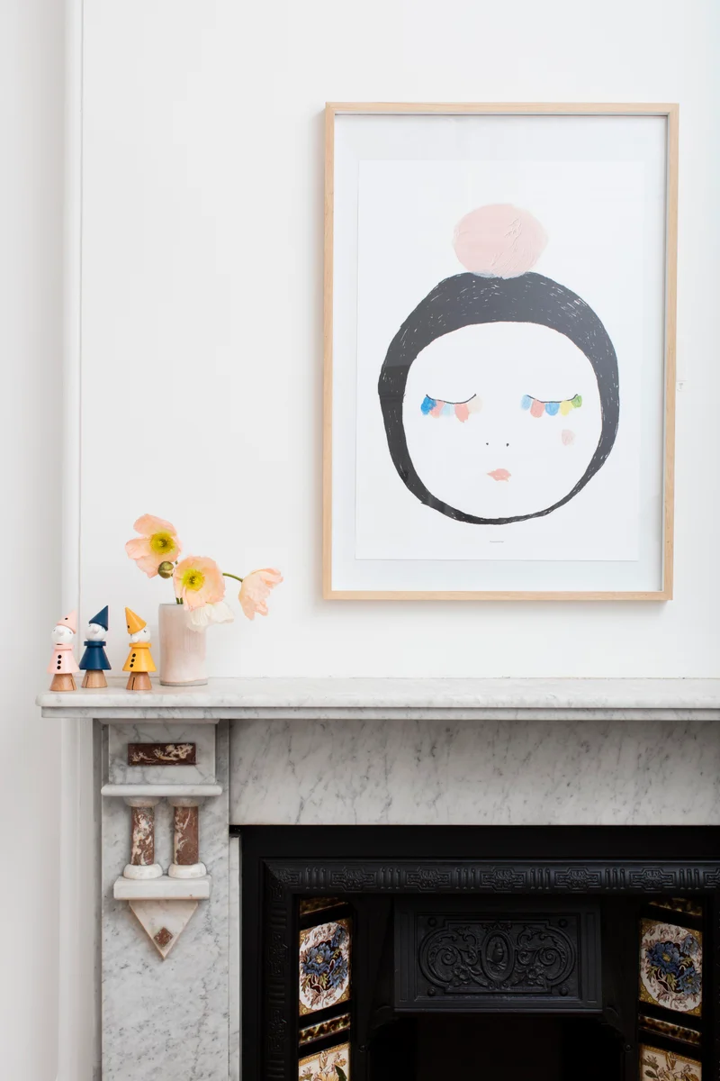 Framed abstract face print displayed on a white wall above a marble mantelpiece with ceramic figurines and flowers.