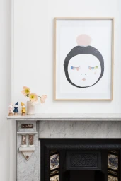 Framed abstract face print displayed on a white wall above a marble mantelpiece with ceramic figurines and flowers.