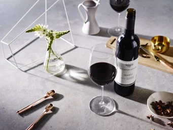 Bottle of red wine next to a filled wine glass on a grey stone tabletop styled with small white flowers in a glass flask.