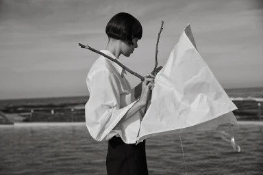 Monochrome image of a woman holding twigs and a white paper structure by the coast.