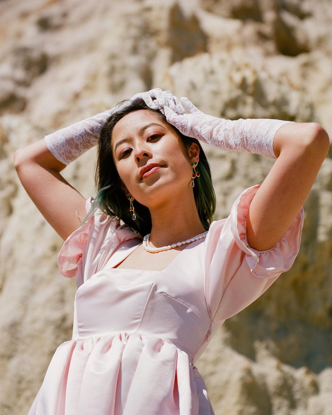Young woman in a light pink dress and white lace gloves poses outdoors against a light-colored rocky cliff backdrop in portrait shot.