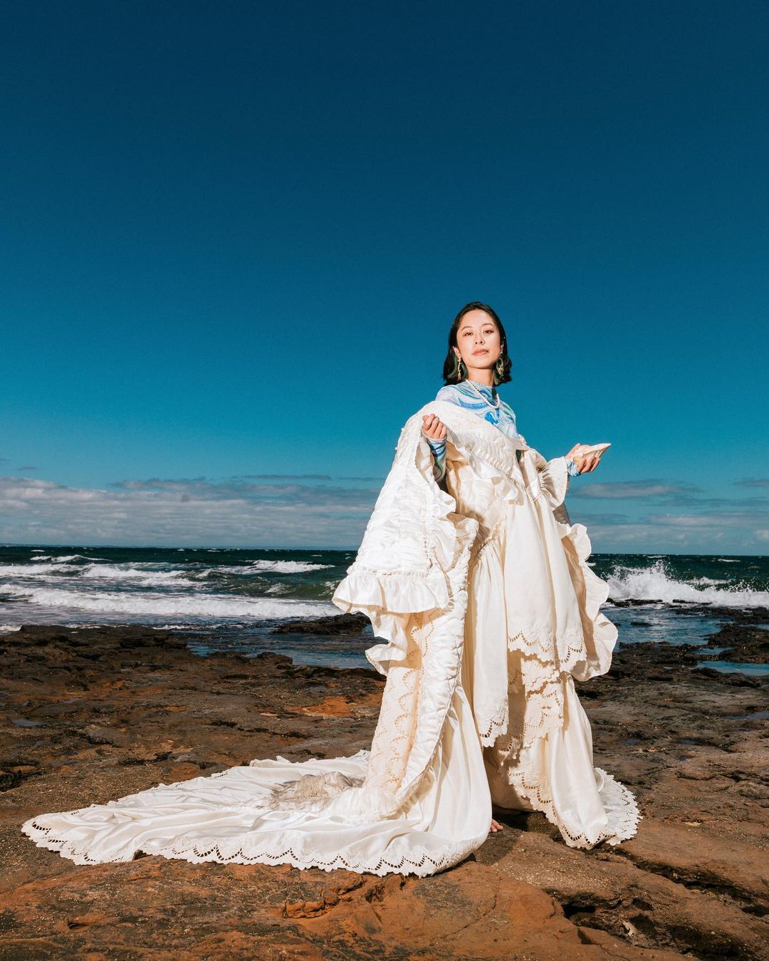 A woman wearing an elaborate white dress stands on a rocky shore with the ocean and blue sky in the background in a fashion editorial.