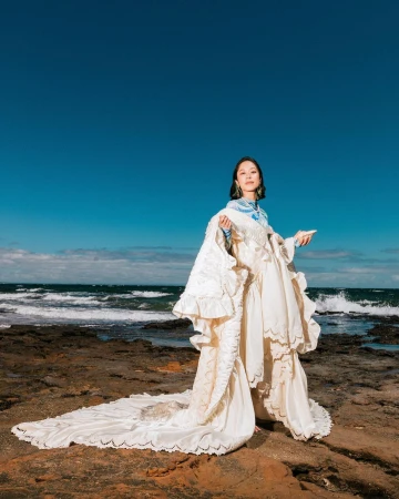 A woman wearing an elaborate white dress stands on a rocky shore with the ocean and blue sky in the background in a fashion editorial.