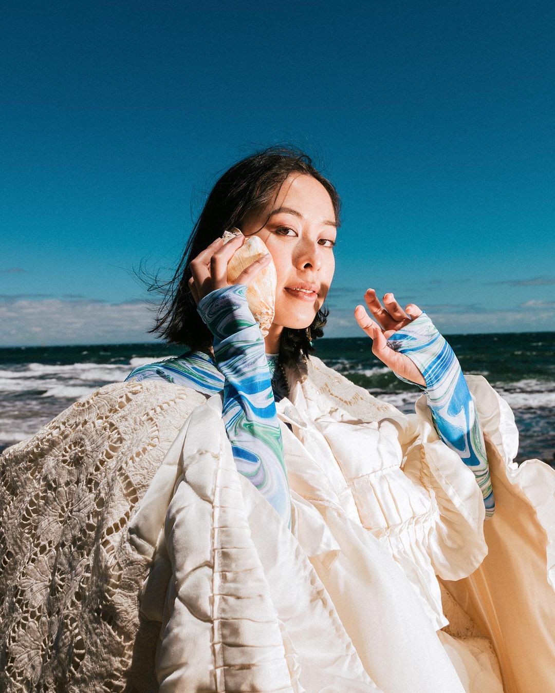 Asian woman poses with a large seashell held to her ear on a beach with blue skies, ocean waves, wearing an ecru colored vintage jacket.