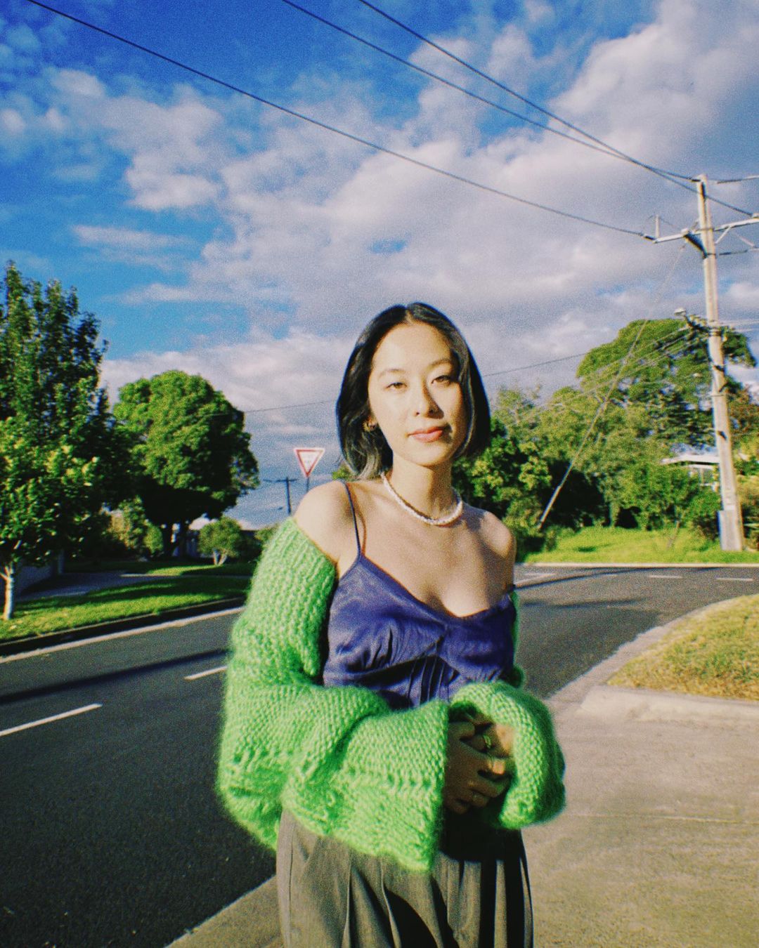 Portrait of a young Asian woman in green sweater and slip dress standing on an asphalt road with blue sky and clouds in the background.
