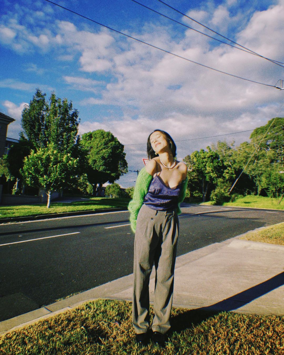 A woman stands outside wearing a blue top, grey pants, and green cardigan, enjoying the sunlight on a street with blue skies.