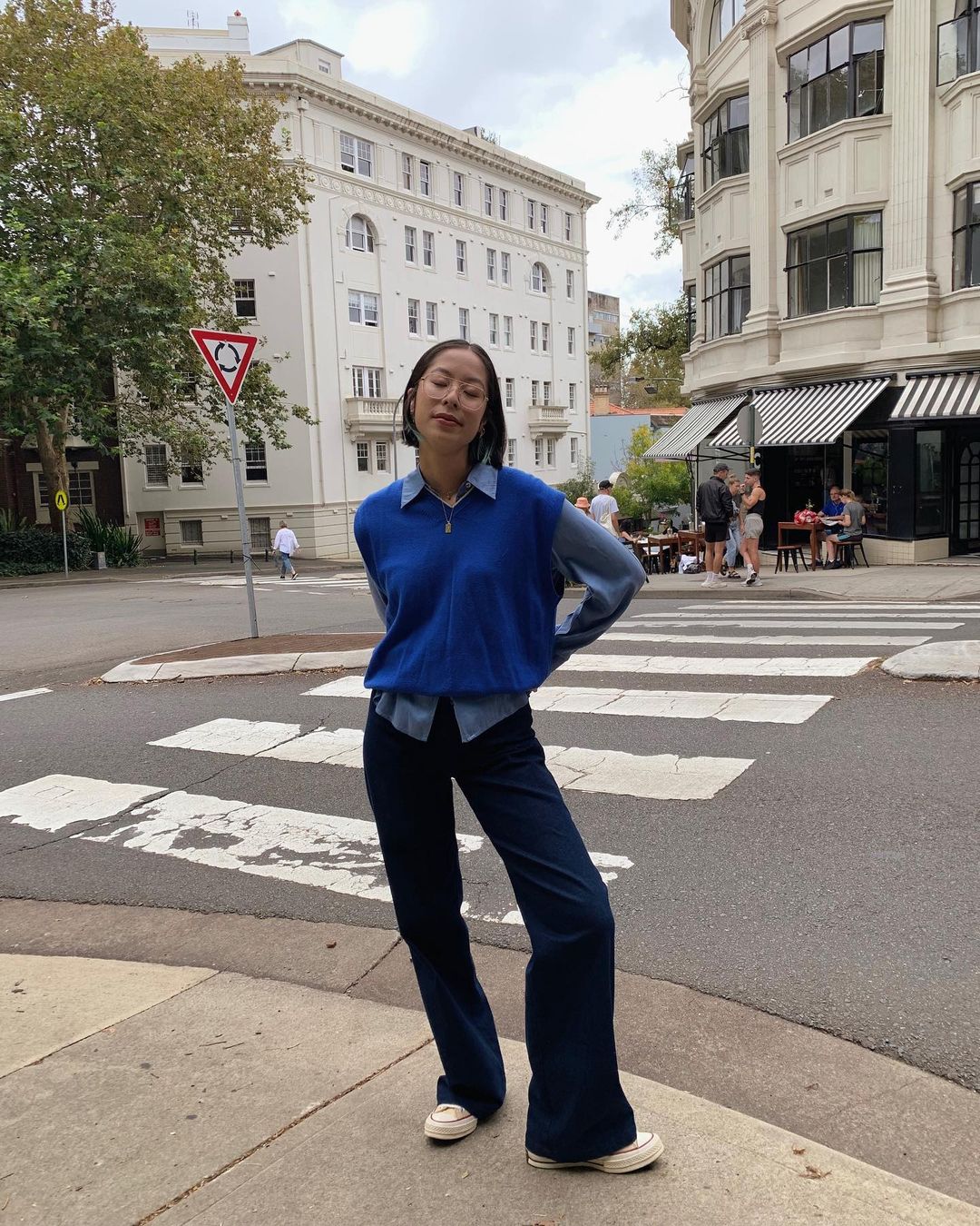 Woman in casual outfit stands on a crosswalk in an urban setting, featuring buildings, street signs, and natural light.
