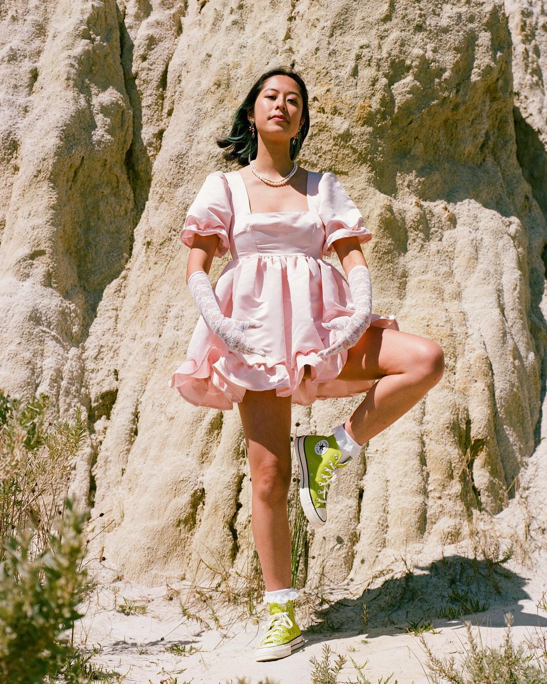 Woman in pink dress and high-top sneakers poses against a rock formation in natural light, creating a whimsical and fashionable outdoor scene.