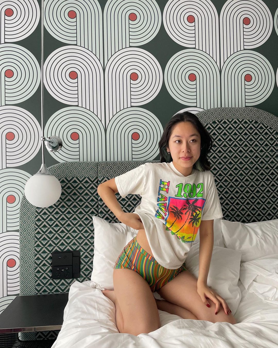 An Asian woman kneels on a bed in a patterned t-shirt and striped shorts, set against a background of geometric wallpaper, inside a bedroom.