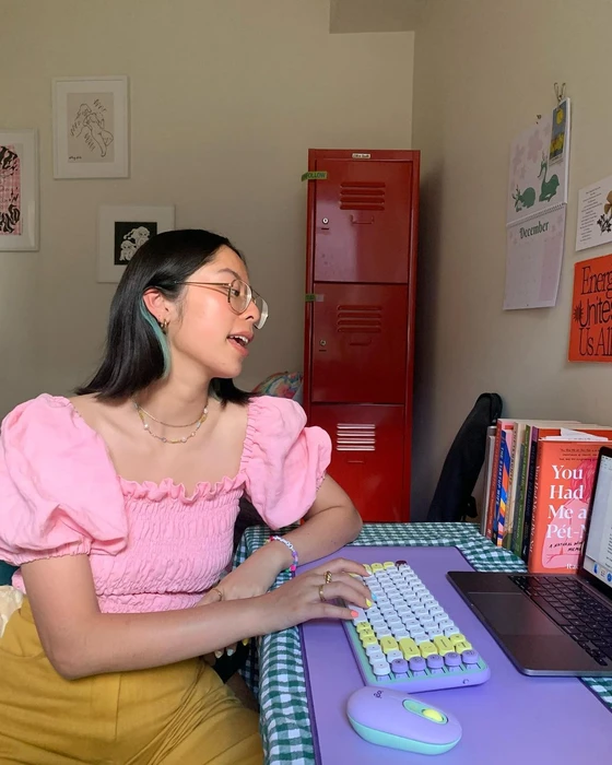 Young east asian woman wearing a pink puff-sleeve top and glasses typing on a pastel keyboard at a desk in a room