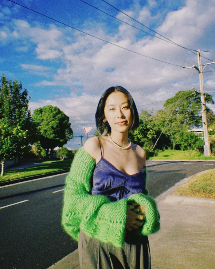 Young woman with dark bobbed hair wearing a bright green knit shrug and blue camisole outdoors on a suburban street.