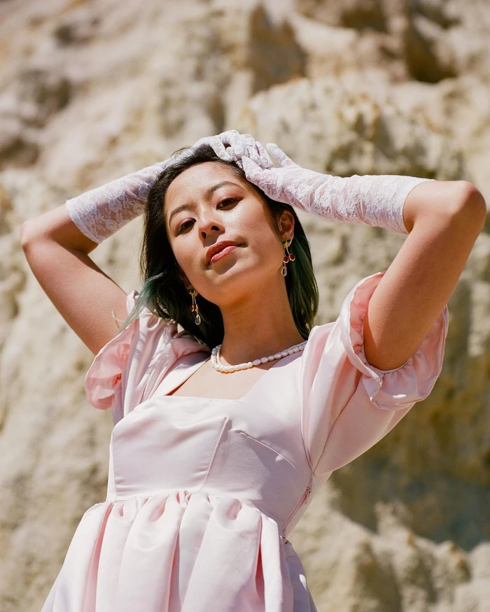 Woman in pink dress with white lace gloves posing against a rocky sandstone cliff background by Maggie Zhou