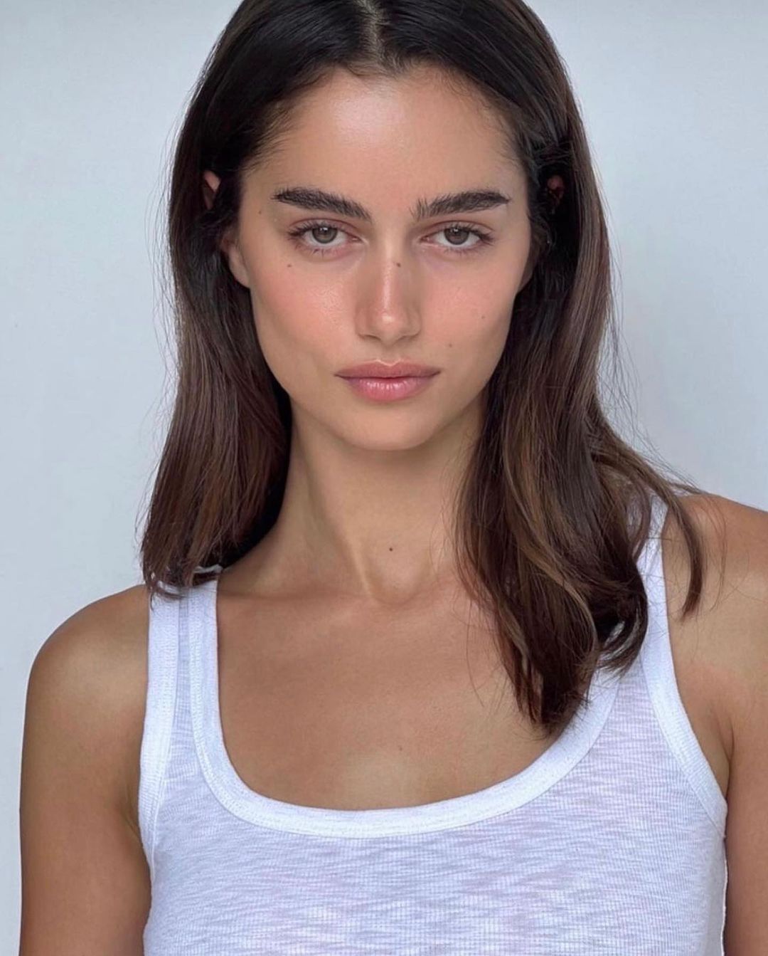 Close-up portrait of a young adult woman with long brown hair and fair skin, wearing a white ribbed tank top against a plain background.