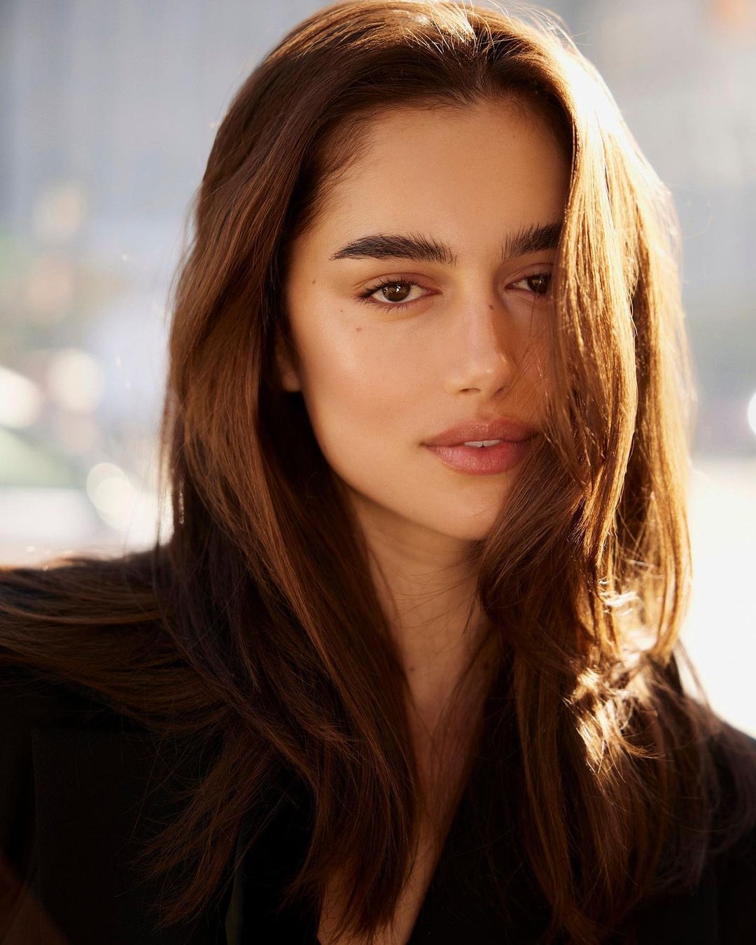 Close-up portrait of an attractive adult woman with brown hair and a black jacket, softly lit in an indoor environment.