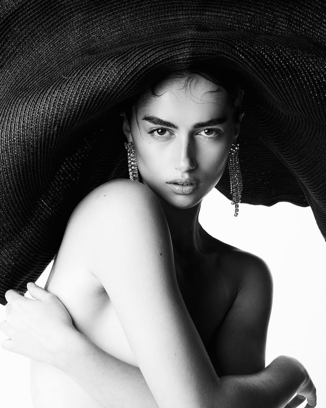 Monochrome portrait of a woman with bare shoulders wearing chandelier earrings and a wide-brimmed hat against a white backdrop.