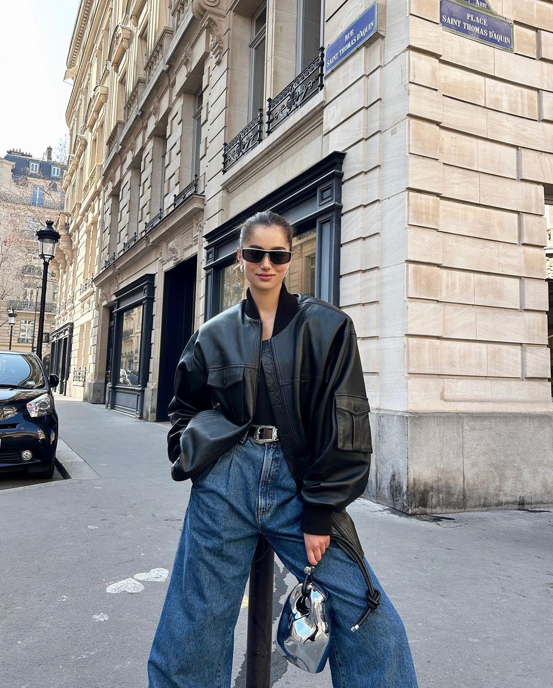 A woman poses on a city street wearing a black leather jacket, blue jeans, and sunglasses with buildings in the background.