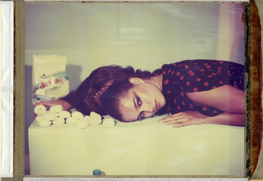 Woman in red and black polka dot dress reclining on a white surface with small white objects and a bag.