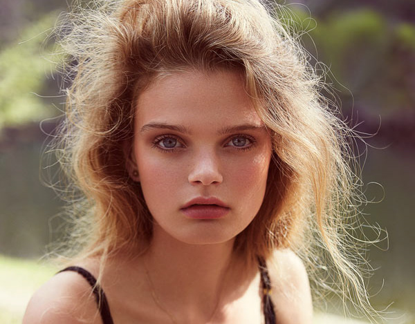 Close-up portrait of a young fair-skinned female model with blonde hair, wearing a black top against a blurred background.