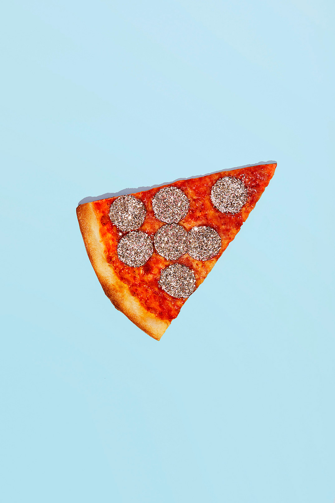 Overhead studio shot of a single slice of pepperoni pizza with glittery toppings against a solid light blue background.