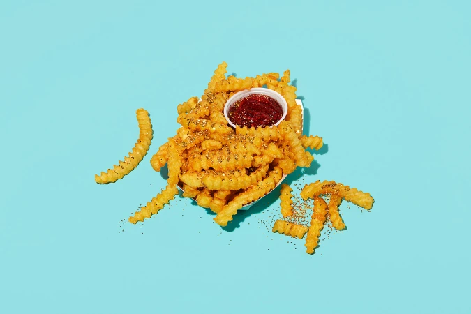 Overhead studio shot of golden crinkle-cut fries with ketchup dipping sauce, sprinkled with black pepper, displayed on a light blue backdrop.