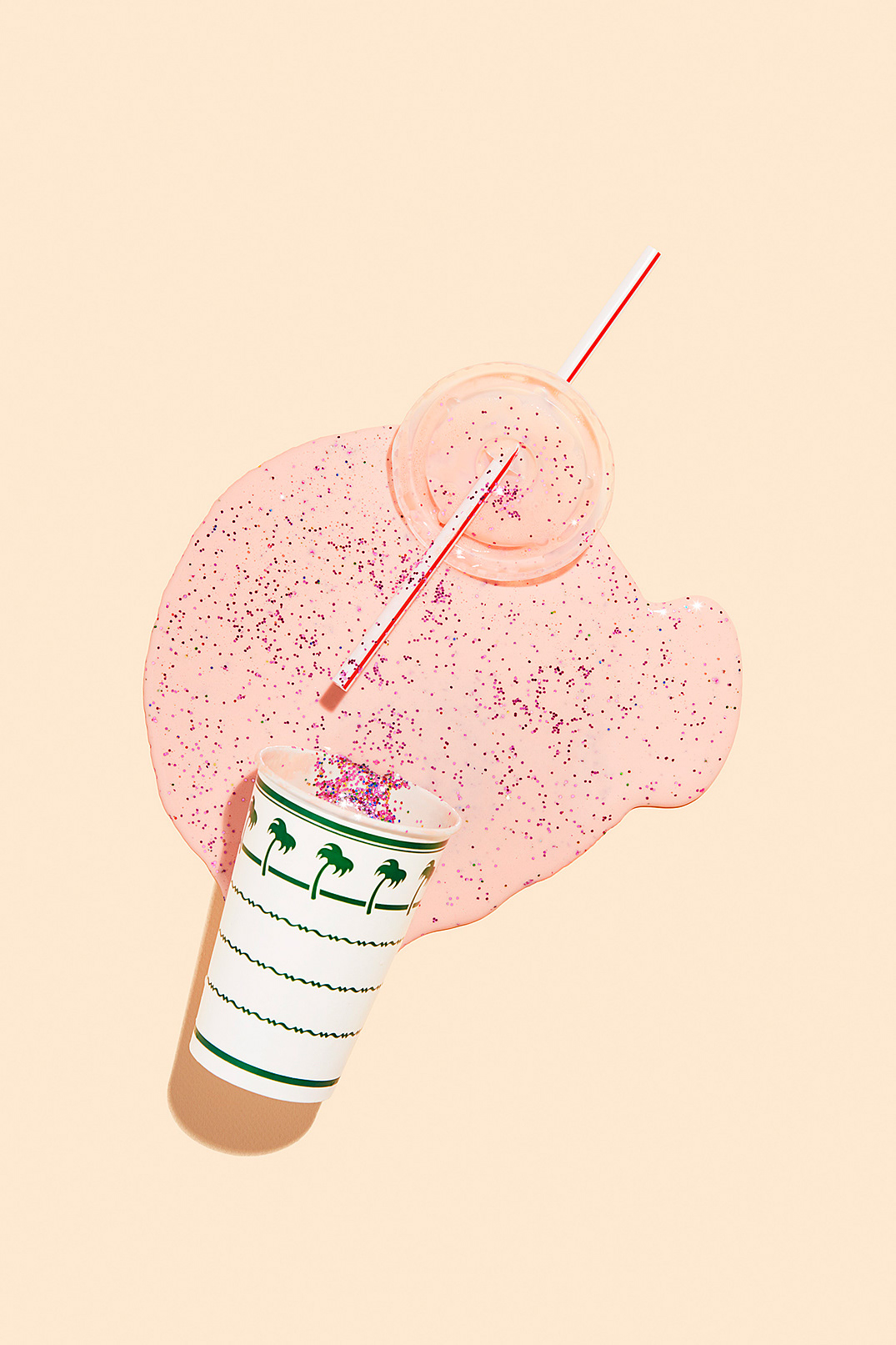 Flat lay photograph of a disposable cup and lid with a straw and spilled pink liquid and glitter on a pale orange background.