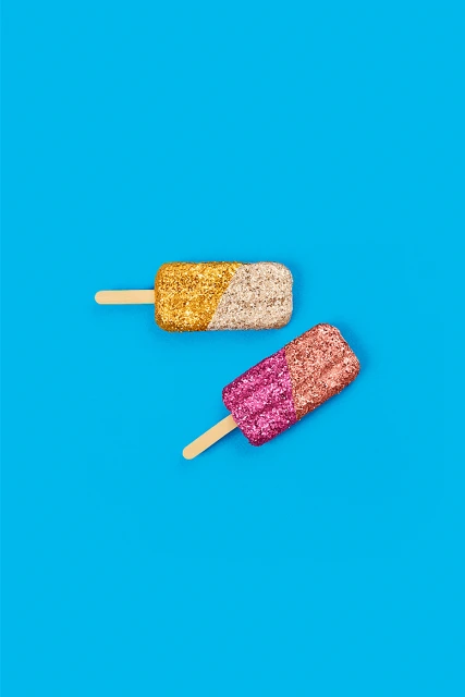 Two colorful glittered ice pops on a vibrant blue background create a fun, eye-catching image perfect for summer or sweet treat themes.