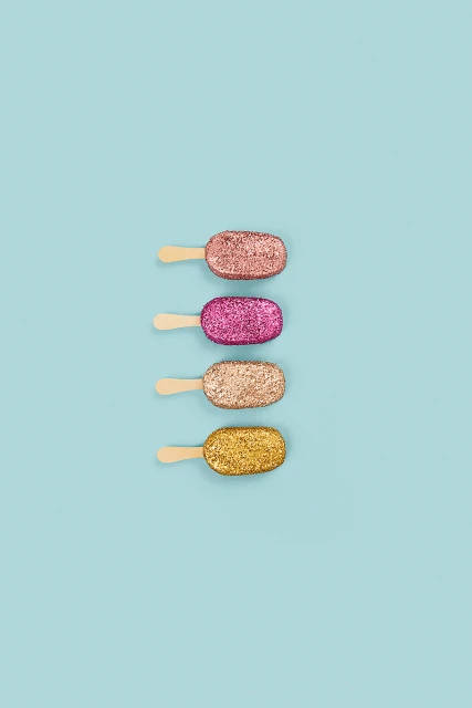 Four glittery dessert popsicles lined up on a light blue background. Shiny pink, gold, and rose-gold colors add to the sweet treat aesthetic.