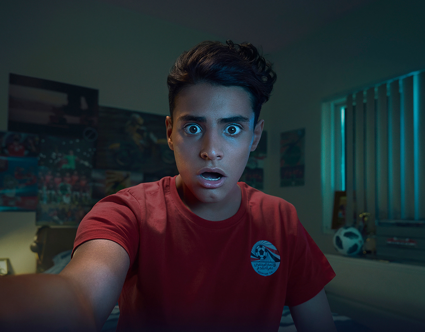 A young man stares wide-eyed at a screen, wearing a red T-shirt in a dimly lit room with posters and a soccer ball.