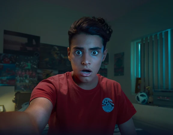 A young man stares wide-eyed at a screen, wearing a red T-shirt in a dimly lit room with posters and a soccer ball.