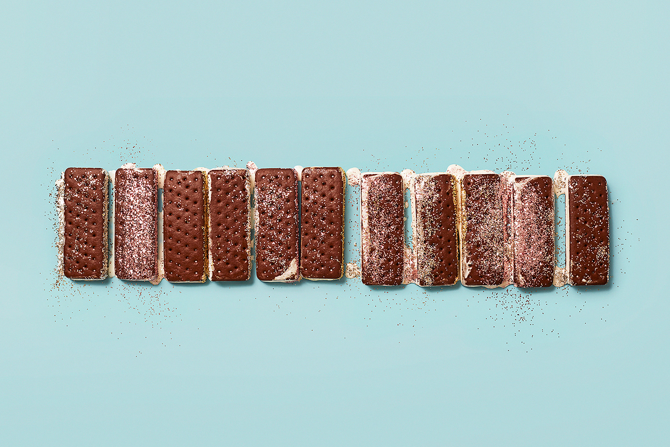 Overhead studio shot of a horizontal row of frosted chocolate ice cream sandwiches lined up on a light blue surface with crumbs.