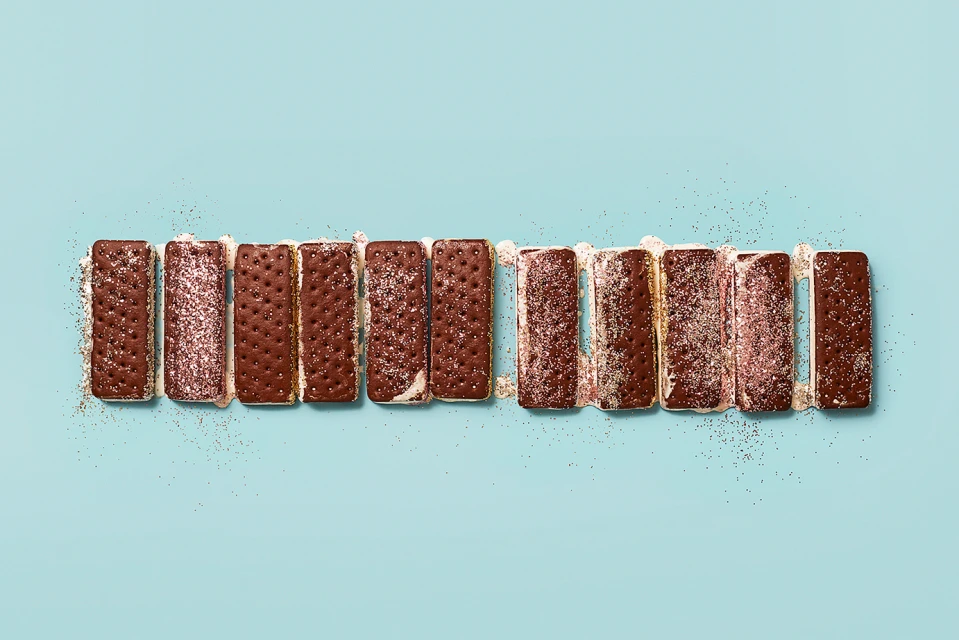 Overhead studio shot of a horizontal row of frosted chocolate ice cream sandwiches lined up on a light blue surface with crumbs.