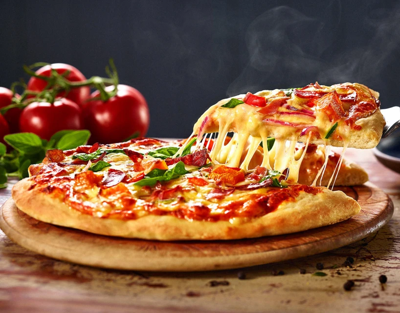 Hot pizza on a wooden board with tomatoes and basil in background, with slice being lifted, showcasing melted cheese.