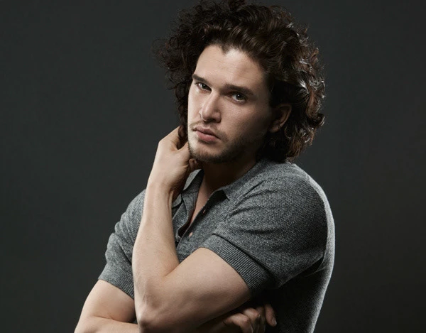 Portrait of a male adult with dark curly hair wearing a grey shirt. He is posing with his hand near his neck, against a dark grey background.
