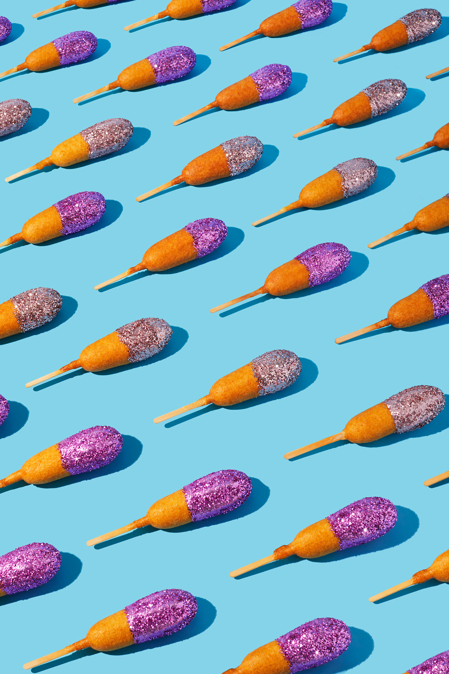 Overhead shot of glitter-covered corn dogs on sticks arranged in a pattern against a solid light blue background with shadow detail.
