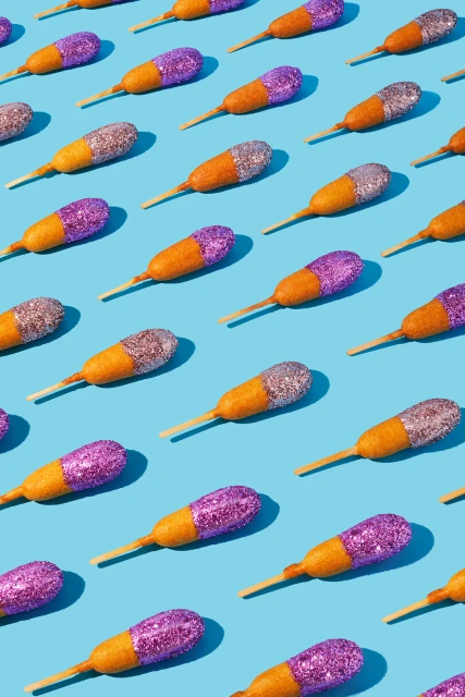 Overhead shot of glitter-covered corn dogs on sticks arranged in a pattern against a solid light blue background with shadow detail.