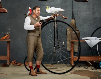 Man in vintage tweed vest and knickerbockers holding a smartphone while leaning on a penny-farthing bicycle with cockatoos.