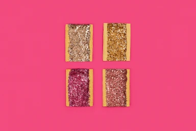 Four rectangular toaster pastries topped with sparkling glitter arranged in a grid on a bright pink background.