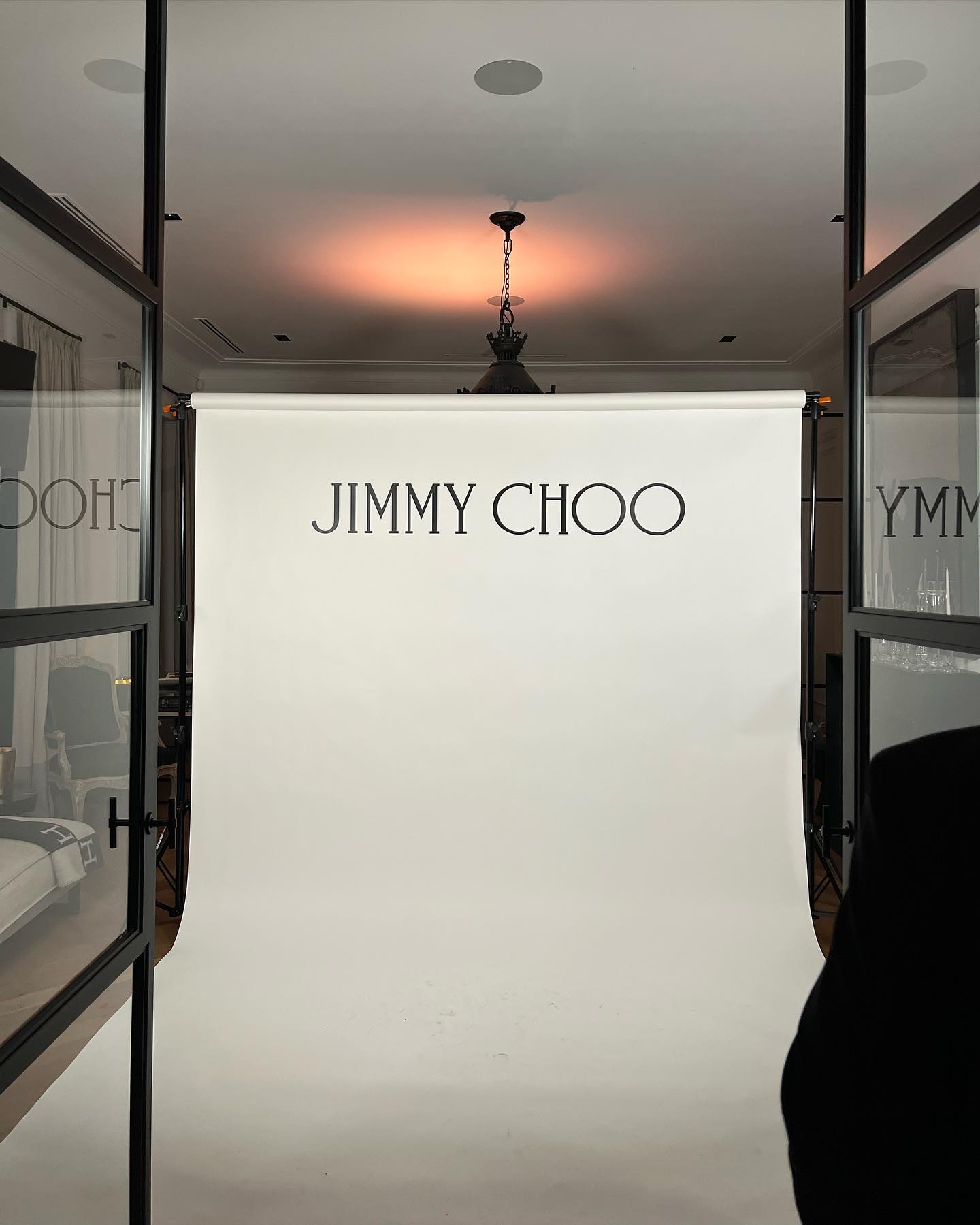 Indoor shot of a white backdrop with the Jimmy Choo logo in black, set up in a room with a black framed door and lighting fixture.
