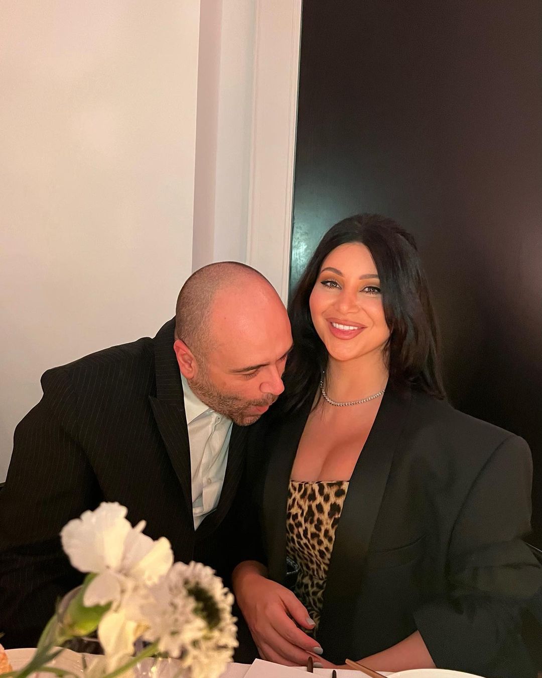 A couple poses indoors; the woman is in a black blazer and animal print dress, while the man wears a black suit and white shirt.