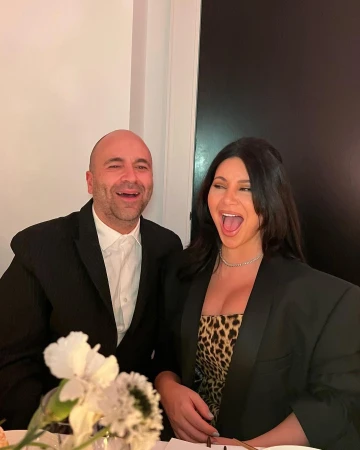 Smiling couple in formal attire poses indoors with a white flower arrangement, creating an elegant and joyful atmosphere.