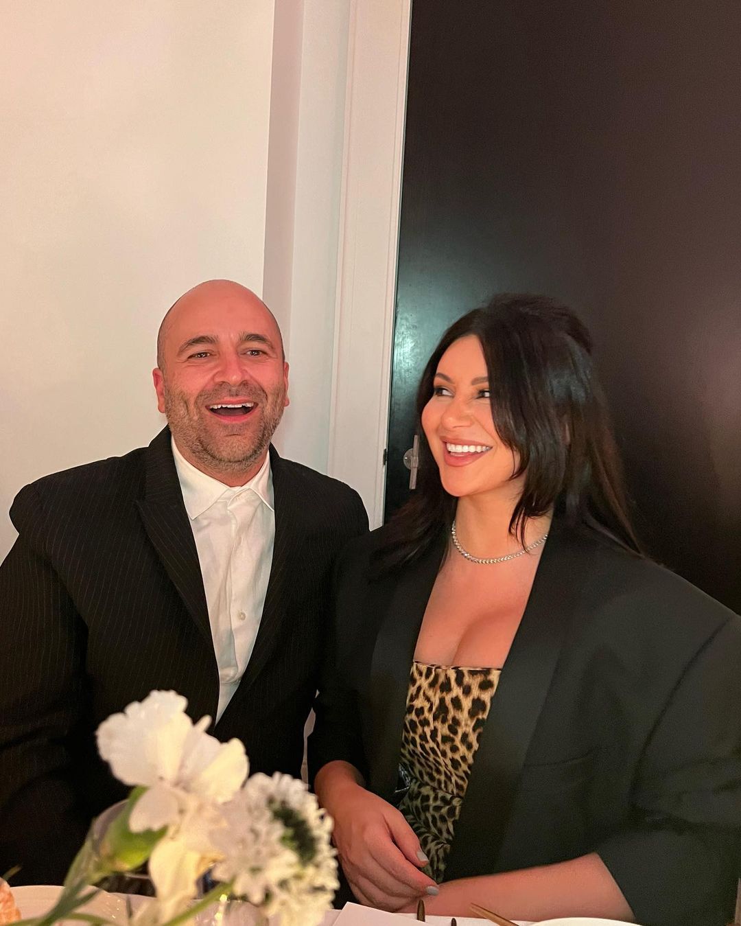 Smiling couple posing indoors, wearing black blazers. The woman wears a leopard print dress. White floral arrangement is on the table.