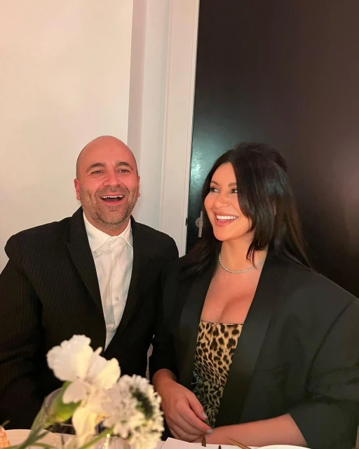 Smiling couple posing indoors, wearing black blazers. The woman wears a leopard print dress. White floral arrangement is on the table.
