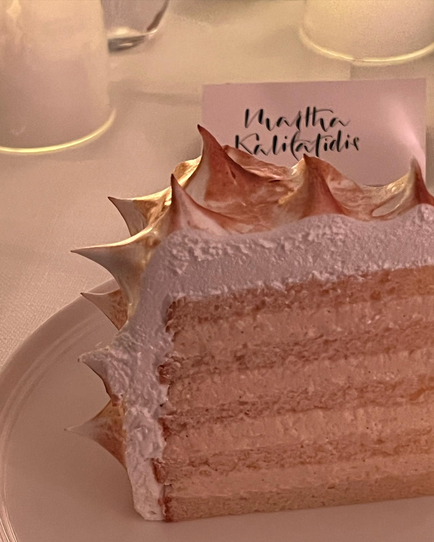 Close-up of a layered cake slice on a white plate with toasted meringue frosting, next to a card on a white tablecloth.