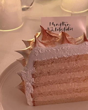 Close-up of a layered cake slice on a white plate with toasted meringue frosting, next to a card on a white tablecloth.
