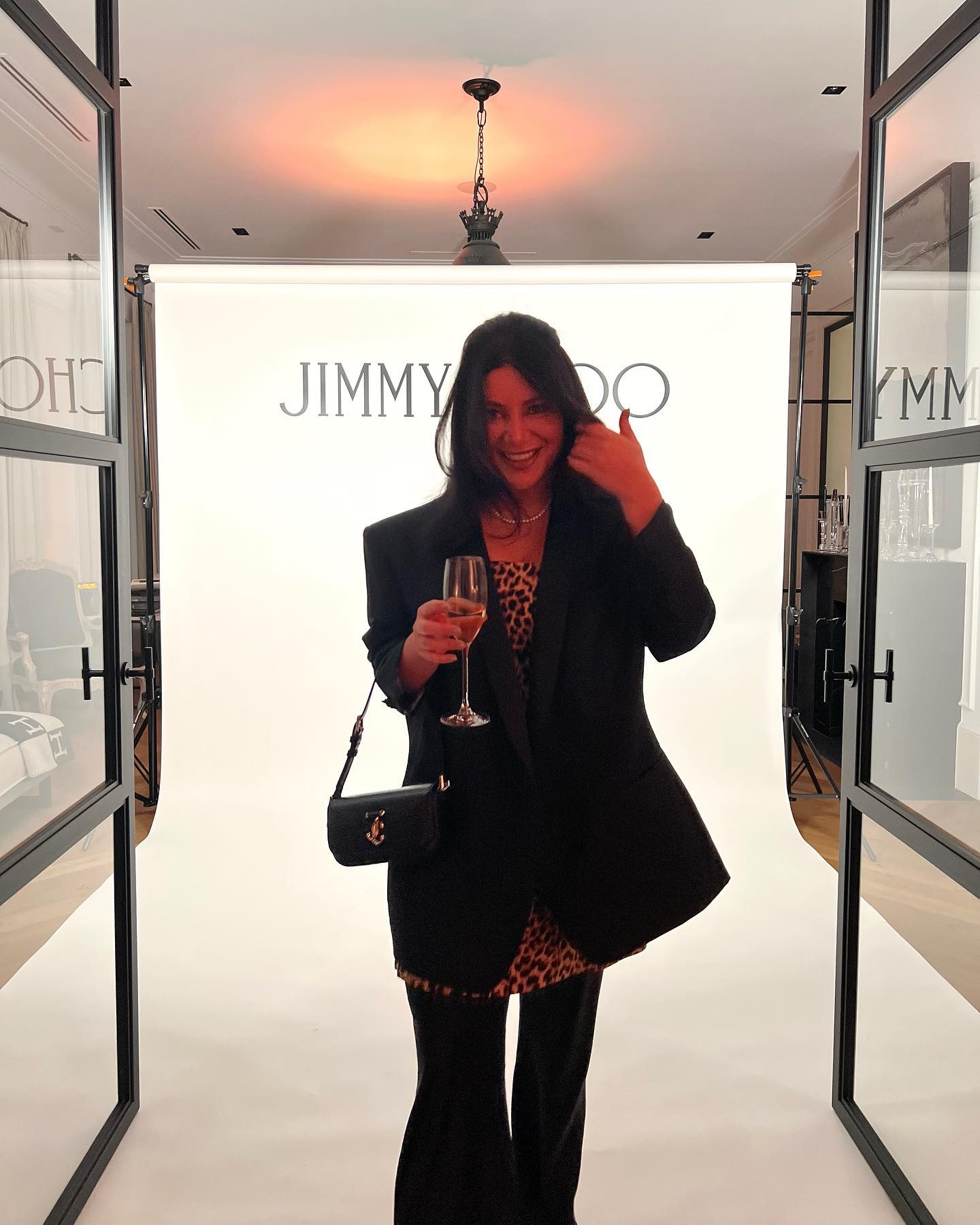 A woman poses indoors at a fashion event wearing a black blazer and holding a champagne glass in front of a Jimmy Choo backdrop.