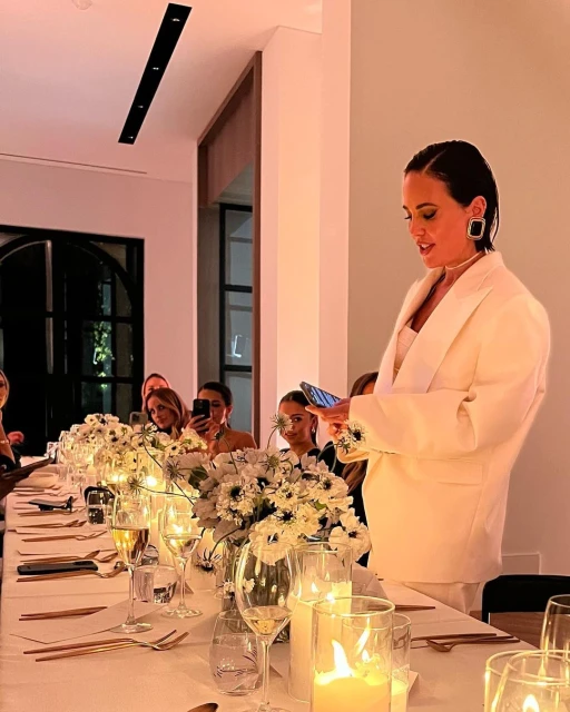 Woman in white suit speaks at a long table decorated with flowers and candles. Guests sit and listen during an event.