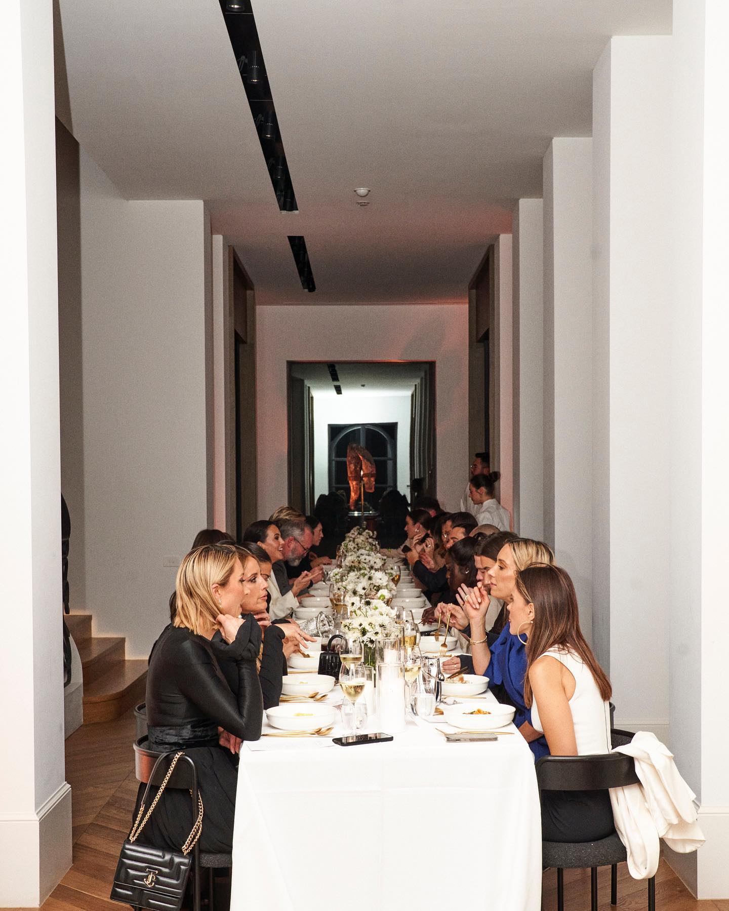 Group of women sitting at long table with white tablecloth in indoor setting. Elegant table setting with floral centerpiece.