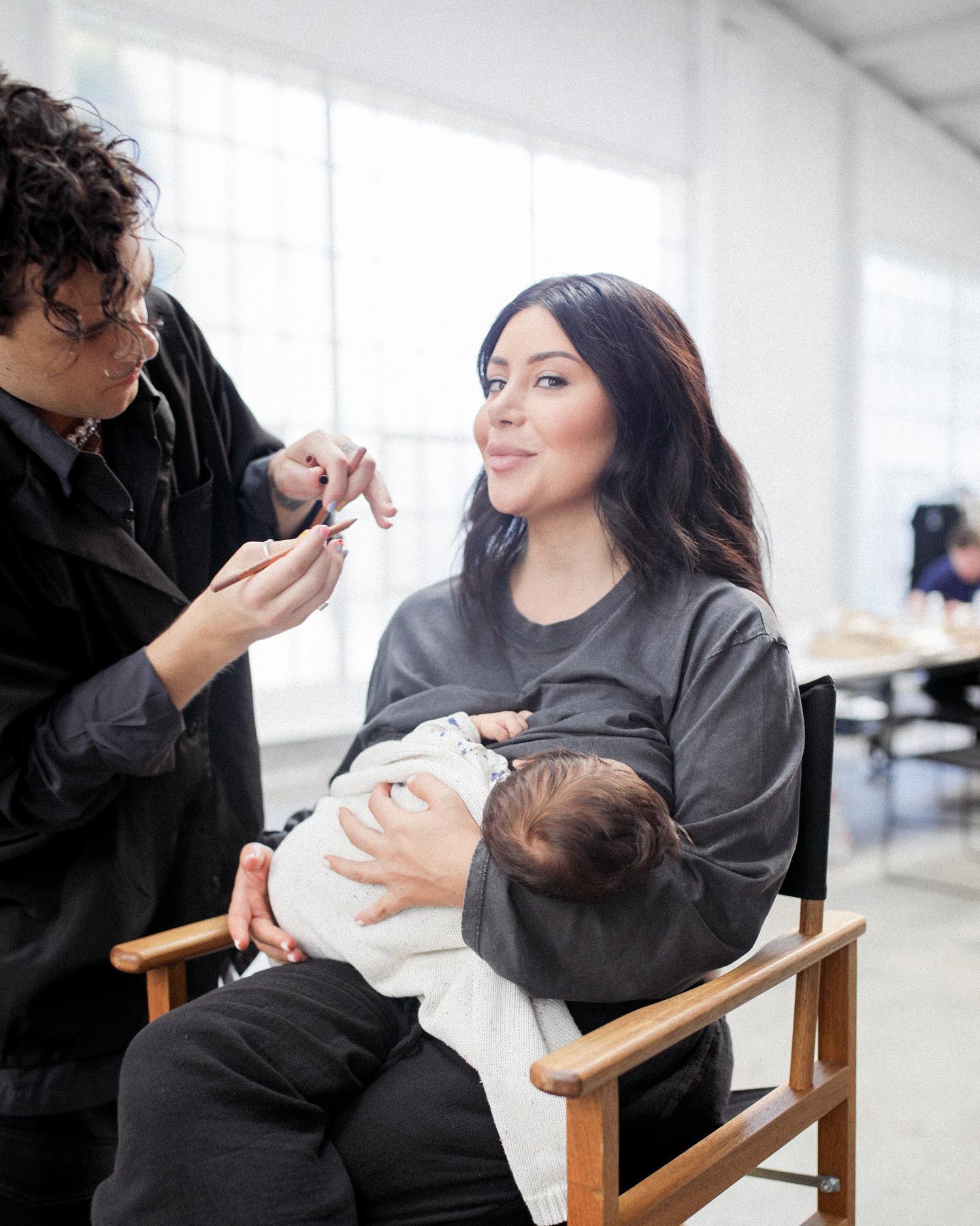 Woman breastfeeding a baby while a makeup artist applies makeup in a bright photo studio with large windows and studio lighting.