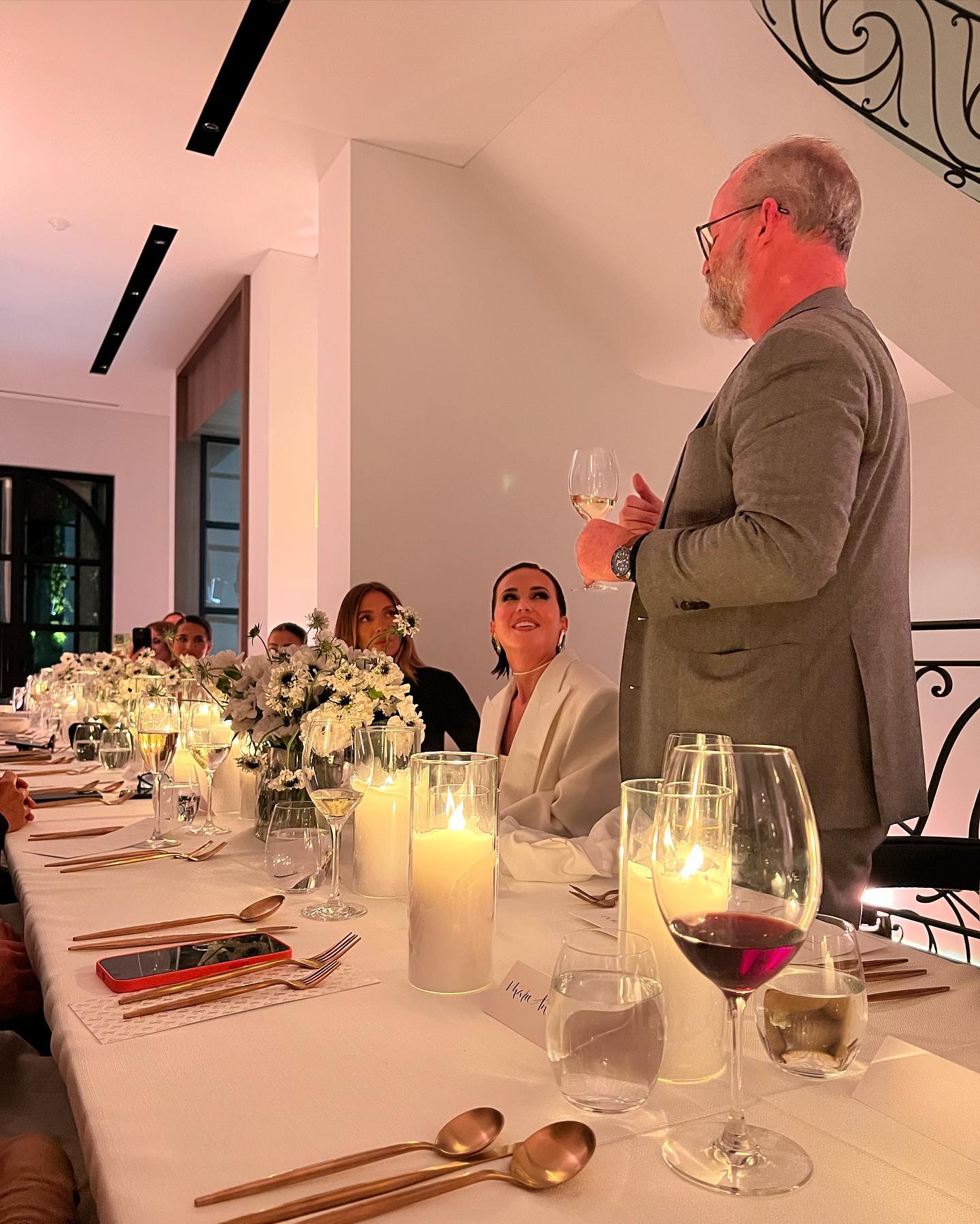 Indoor gathering at a long table with an adult male giving a toast, surrounded by dinnerware, candles, and floral arrangements.
